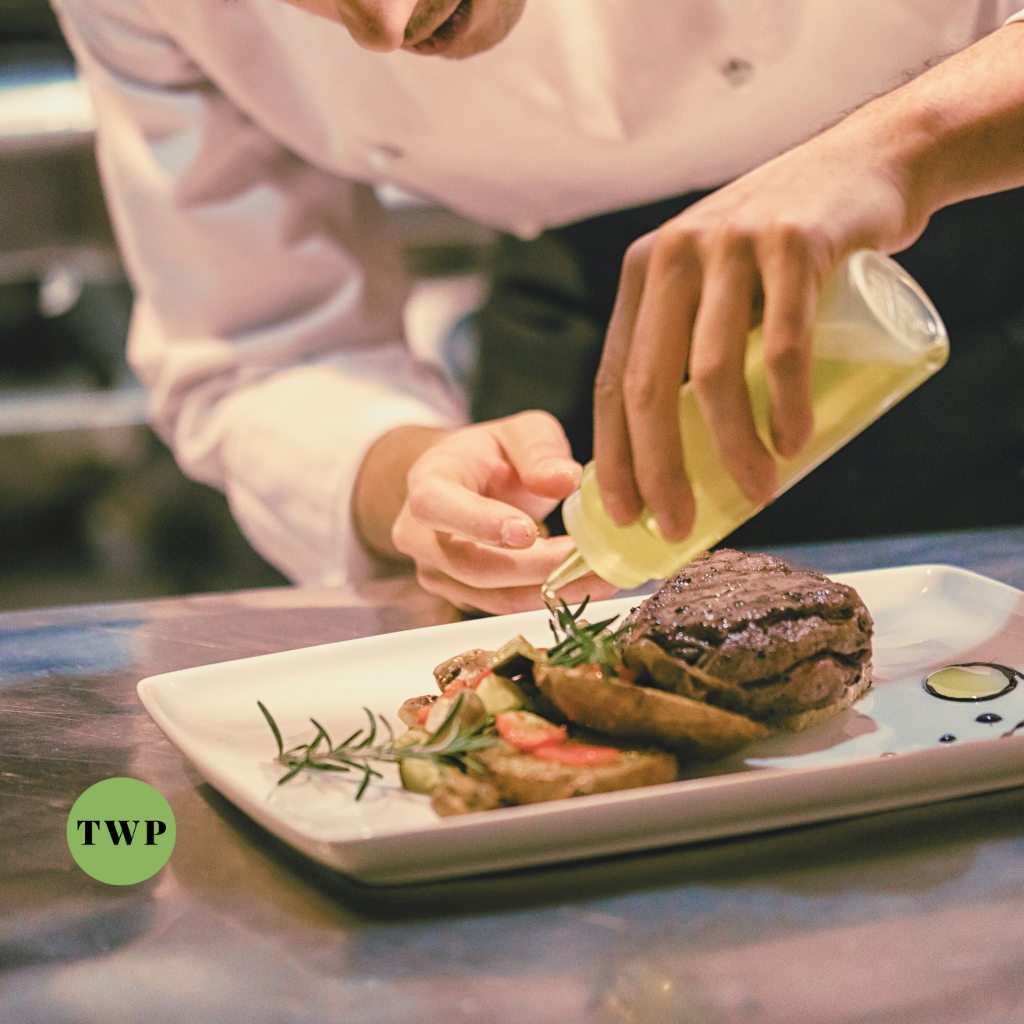 Chef in a professional kitchen meticulously garnishing a steak dish with a drizzle of sauce, showcasing culinary expertise and attention to detail, complemented by the TWP logo.